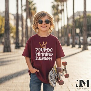Può includere: T-shirt bordeaux con la scritta bianca "YOUR WINNING DESIGN" e una grafica a corona dorata. La persona indossa occhiali da sole e tiene in mano uno skateboard. La persona indossa jeans blu.