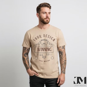 Könnte beinhalten: Ein beigefarbenes T-Shirt mit Rundhalsausschnitt und dem Text "YOUR DESIGN WINNING JUST MOCKS" in einem dekorativen Design. Das T-Shirt wird von einer Person mit Tätowierungen an den Armen getragen.
