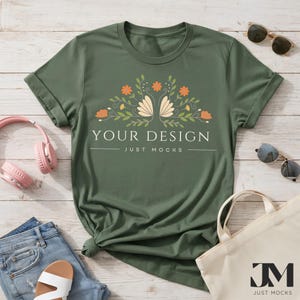 Könnte beinhalten: Ein olivgrünes T-Shirt mit hochgekrempelten Ärmeln, mit einem floralen Design und dem Text "YOUR DESIGN JUST MOCKS". Das Shirt ist mit Sonnenbrille, Tragetasche, Kopfhörern und Jeansshorts auf einer Holzoberfläche gestylt.