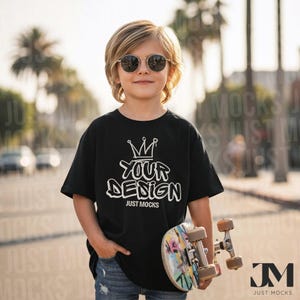 Puede incluir: Una camiseta negra con el texto blanco "YOUR DESIGN", con un gráfico de corona. La camiseta se combina con vaqueros azules. Se sostiene en la mano una patineta con ilustraciones coloridas. El fondo incluye palmeras y una escena callejera borrosa.