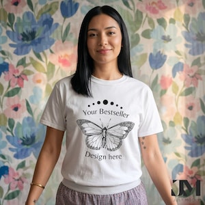Può includere: T-shirt bianco con grafica a farfalla nera e la scritta "Your Bestseller Design here". La maglietta a maniche corte ha lo scollo tondo. Lo sfondo è un motivo floreale con fiori rosa e blu.