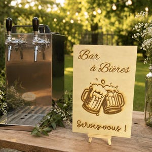 Peut inclure: Un distributeur de bière en acier inoxydable à deux becs à côté d'un panneau en bois rectangulaire sur un petit chevalet. Le panneau porte l'inscription gravée Bar à Bières et Servez-vous ! au-dessus de deux chopes de bière moussantes.