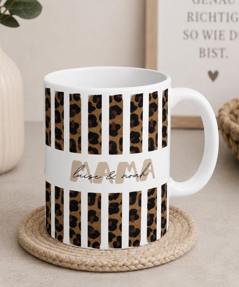 May include: A white ceramic mug featuring a repeating pattern of vertical leopard print stripes. A horizontal white band across the center displays the word MAMA in large beige letters with the names luise & noah written in black script below.