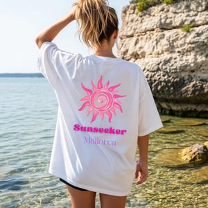 May include: A white t-shirt featuring a pink graphic of a stylized sun on the back. Below the sun, the text reads Sunseeker in bold pink letters and Mallorca in smaller purple letters. The shirt has short sleeves and a loose, relaxed fit.