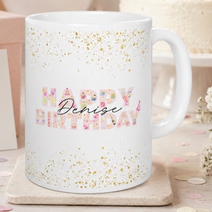 May include: A white ceramic mug featuring a gold glitter pattern at the top and bottom. The center displays the text "HAPPY BIRTHDAY" in light pink block letters filled with small party icons, with the name "Denise" written in black script.