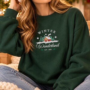 May include: A dark green crewneck sweatshirt featuring an embroidered graphic of a small cabin in a snowy forest. The text reads Winter Wonderland and EST 2024. The design includes white snowflakes and green pine trees, with a red cabin roof.