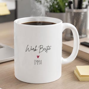 May include: A white ceramic mug filled with dark coffee and steam rising from the top. The front of the mug features the black text "Work Bestie" above a small red heart and the name "EMMA" printed in a simple, clean, black sans-serif font.