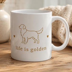 May include: A white ceramic mug featuring a gold-colored line art illustration of a Golden Retriever dog. The design includes a small heart on each side of the dog and the text "life is golden" printed in gold lettering below the illustration.