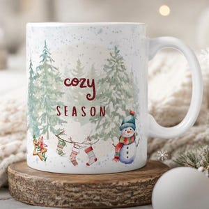 May include: A white ceramic mug featuring a watercolor illustration of green pine trees, a snowman wearing a blue hat and red scarf, and a string of hanging stockings. The text "cozy SEASON" is printed in dark red in the center of the mug.