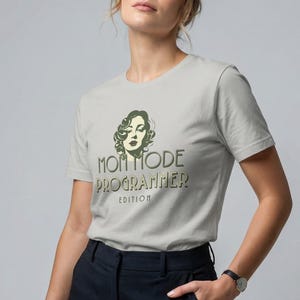 May include: A pale grey t-shirt featuring the text "MOM MODE PROGRAMMER EDITION" and a woman's face illustration. The t-shirt is worn with dark blue trousers.