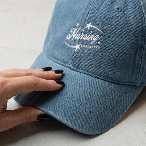 May include: A light blue denim baseball cap with the word "Nursing" in white script, arched above the word "Graduated". The cap has a curved brim and a six-panel design. The text is surrounded by stars.