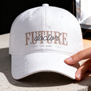 May include: White baseball cap with the text "FUTURE doctor" in a brown and black font. The text "EST. 2026" is below the main text. The cap is on a light gray surface.
