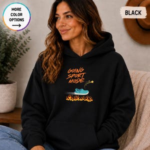 May include: A black hooded sweatshirt featuring a graphic print with the orange text "GOING SPORT MODE" above a light blue clog shoe moving quickly with a trail of fire and a small yellow star icon positioned below the shoe graphic.