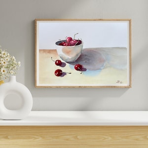 Könnte beinhalten: Aquarellmalerei einer Schüssel voller reifer Kirschen, mit mehreren Kirschen verstreut. Die Schüssel ist weiß mit einem rosafarbenen Farbton, und die Kirschen sind tiefrot. Das Gemälde ist in einem hellen Holzrahmen gerahmt.