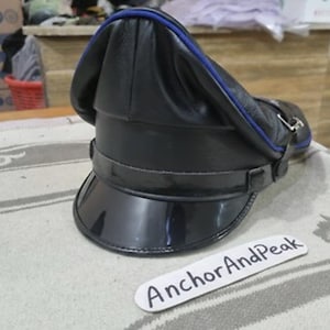 May include: A black leather hat with a shiny, curved visor and a blue trim. The hat has a structured design with a strap and buckle detail. The text "AnchorAndPeak" is on a white label in the foreground.