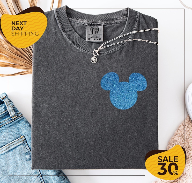 a gray t-shirt with a blue glittery Mickey Mouse design, along with a pair of blue jeans and a beige sweater. The t-shirt is displayed on a white background, and there are yellow and black sale signs indicating a 30% discount.