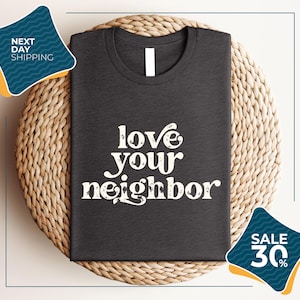 a gray t-shirt with the text "love your neighbor" printed on it, displayed on a woven placemat. The t-shirt is placed on a round wicker basket, and there are two sale signs in the image.
