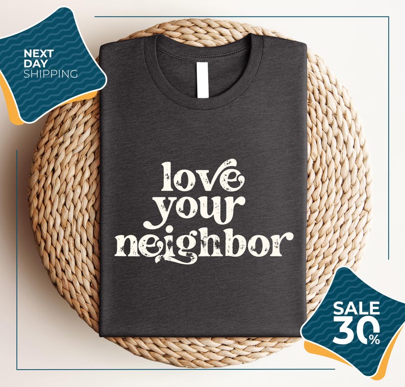 a gray t-shirt with the text "love your neighbor" printed on it, displayed on a woven placemat. The t-shirt is placed on a round wicker basket, and there are two sale signs in the image.