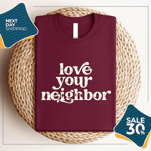 a burgundy t-shirt with the text "love your neighbor" printed on it, displayed on a woven placemat. The t-shirt is placed on a round wicker basket, and there are two sale signs in the image, one indicating a 30% discount and the other promoting next day shipping.