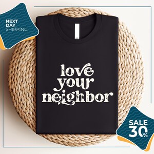 a black t-shirt with the text "love your neighbor" printed on it, displayed on a woven placemat. The t-shirt is placed on a round wicker basket, and there are two sale signs in the image.