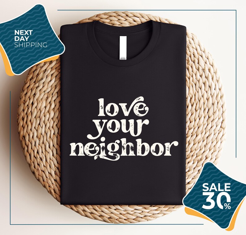 a black t-shirt with the text "love your neighbor" printed on it, displayed on a woven placemat. The t-shirt is placed on a round wicker basket, and there are two sale signs in the image.