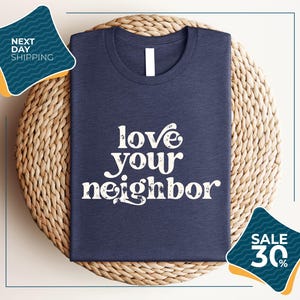 A navy blue t-shirt with the text "love your neighbor" printed on it, displayed on a woven placemat. The t-shirt is folded and placed on a round wicker placemat. There are two sale signs in the image, one on the left side and one on the right side, indicating a sale or discount.