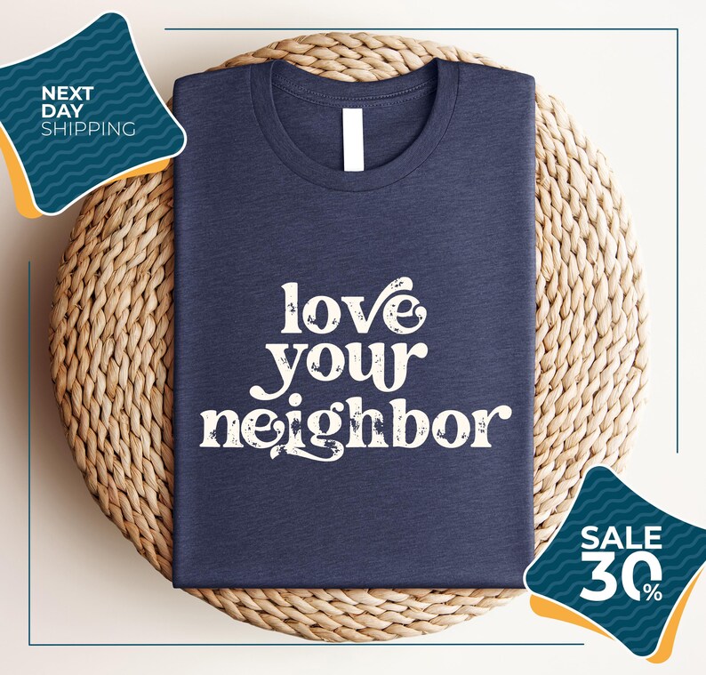 A navy blue t-shirt with the text "love your neighbor" printed on it, displayed on a woven placemat. The t-shirt is folded and placed on a round wicker placemat. There are two sale signs in the image, one on the left side and one on the right side, indicating a sale or discount.