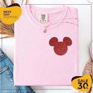 a pink t-shirt with a red glitter Mickey Mouse logo, a necklace, and a pair of blue jeans, all displayed on a white background.