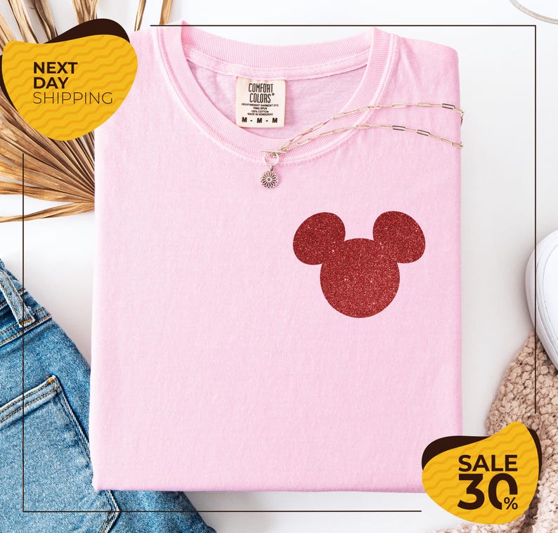 a pink t-shirt with a red glitter Mickey Mouse logo, a necklace, and a pair of blue jeans, all displayed on a white background.
