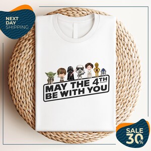 A white t-shirt with a graphic design featuring characters from the Star Wars franchise, along with the text "May the 4th be with you".