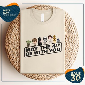 A beige t-shirt with a graphic design featuring five characters from the Star Wars franchise, including Yoda, Darth Vader, Princess Leia, and Chewbacca, along with the text "May the 4th be with you".