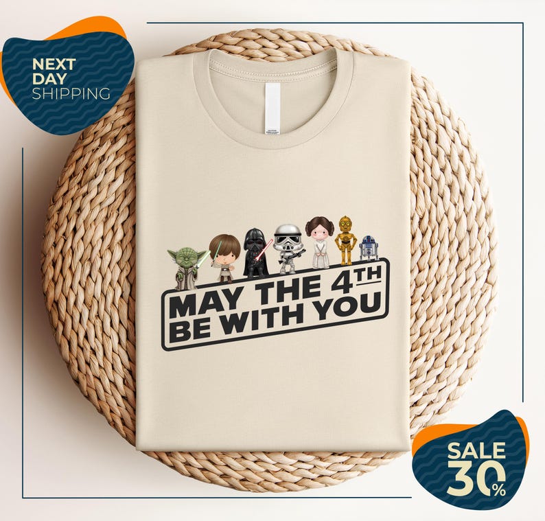 A beige t-shirt with a graphic design featuring five characters from the Star Wars franchise, including Yoda, Darth Vader, Princess Leia, and Chewbacca, along with the text "May the 4th be with you".