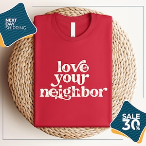a red t-shirt with the text "love your neighbor" printed on it, displayed on a woven placemat. The t-shirt is folded and placed on a round wicker basket. There are two sale signs in the image, one indicating a 30% discount and the other promoting next day shipping.