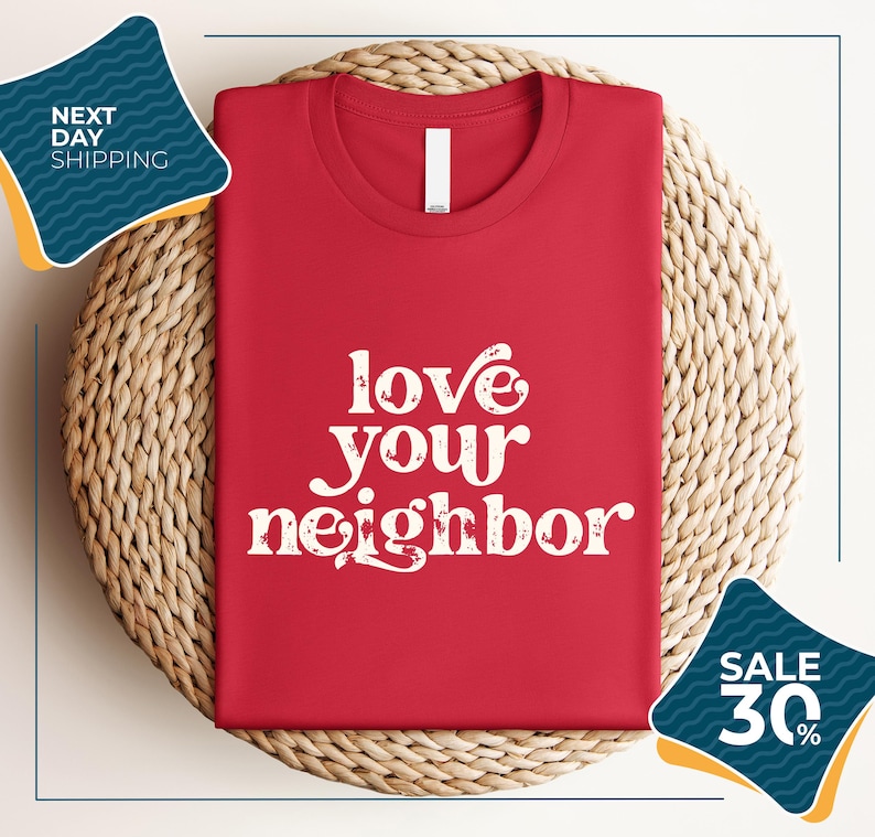 a red t-shirt with the text "love your neighbor" printed on it, displayed on a woven placemat. The t-shirt is folded and placed on a round wicker basket. There are two sale signs in the image, one indicating a 30% discount and the other promoting next day shipping.