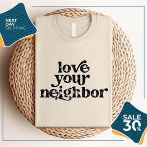 a beige t-shirt with the text "LOVE YOUR NEIGHBOR" printed on it, displayed on a woven basket or mat. There are two sale signs in the image, one on the left side and one on the right side, indicating a sale or discount on the t-shirt.