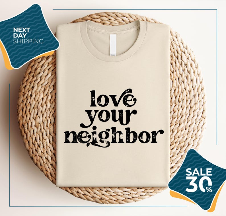 a beige t-shirt with the text "LOVE YOUR NEIGHBOR" printed on it, displayed on a woven basket or mat. There are two sale signs in the image, one on the left side and one on the right side, indicating a sale or discount on the t-shirt.