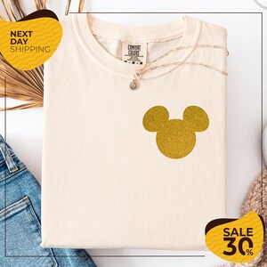 a white t-shirt with a gold glitter Mickey Mouse design, placed on a white background. There are yellow and black sale signs in the image, indicating a sale or discount.