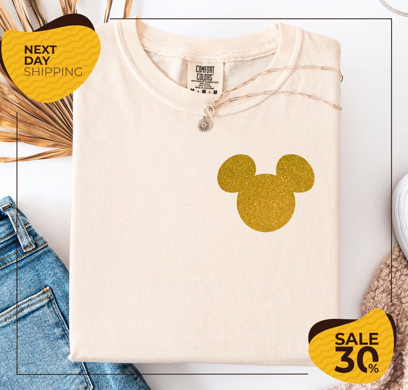 a white t-shirt with a gold glitter Mickey Mouse design, placed on a white background. There are yellow and black sale signs in the image, indicating a sale or discount.