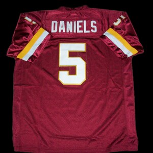 Pode incluir: Camisola de futebol bordô com o nome "DANIELS" e o número "5" em branco e dourado. As mangas têm listras brancas e douradas. A camisola é feita de um material de malha respirável.