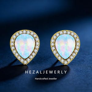 May include: A pair of gold-toned teardrop earrings. Each earring features a pear-shaped opal center stone surrounded by a halo of small, clear crystals. The earrings are set against a dark blue background. The text "HEZALJEWERLY" and "Handcrafted Jeweller" are visible.