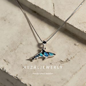 May include: A silver shark pendant necklace with blue and black accents. The shark pendant has a detailed design with blue opal-like stones. The necklace is displayed on a textured, light-colored surface. The text "HEZALJEWERLY" and "Handcrafted Jeweller" are visible.