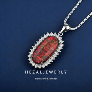 May include: A silver pendant necklace featuring an oval-shaped, fiery red opal gemstone. The opal is surrounded by a halo of small, clear gemstones. The necklace has a delicate silver chain. The text "HEZALJEWERLY" and "Handcrafted Jeweller" are visible.