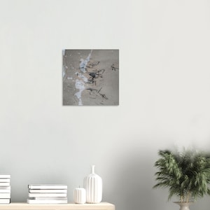 May include: Abstract artwork on a square canvas. The piece features a textured, light grey background with scattered black lines and white paint splatters. The artwork is mounted on a white wall, with a neutral-toned shelf and decorative vases in the foreground.