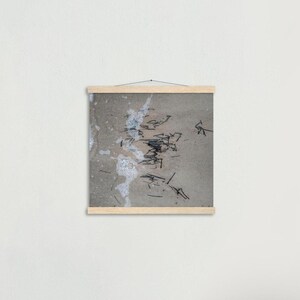 May include: A framed print showing a close-up of a sandy beach with dark debris and white foam. The print is mounted with a natural wood frame, creating a minimalist aesthetic. The artwork captures a moment of the ocean's edge.