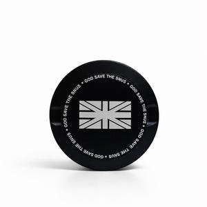 Può includere: Un contenitore nero e rotondo con la scritta "GOD SAVE THE SNUS" sul bordo. Il centro presenta un disegno della bandiera Union Jack bianca.