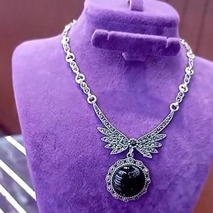 Peut inclure: Un collier argenté avec un pendentif noir est présenté sur un buste en velours violet. Le collier présente un motif en forme d'ailes avec des accents verts et un pendentif circulaire en pierre noire. La chaîne a un design délicat.