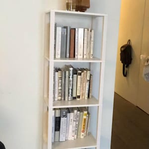May include: A white, four-shelf bookcase filled with books of various sizes and colors. A decorative wooden vase with dried flowers sits on top. The bookcase is against a light blue wall.
