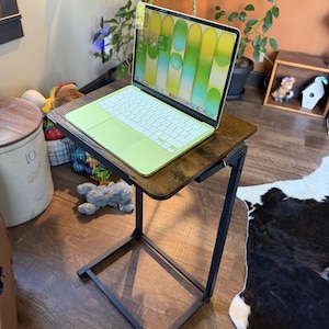 May include: A laptop with a light green keyboard sits on a brown, wood-grain side table with a black metal frame. The laptop screen displays a colorful, abstract design. The table is designed to slide under a couch or chair.
