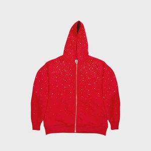 May include: A red zip-up hoodie with a hood, adorned with scattered gold-colored embellishments. The hoodie features a full-length zipper and is made of a soft material. The garment is displayed against a plain white background.