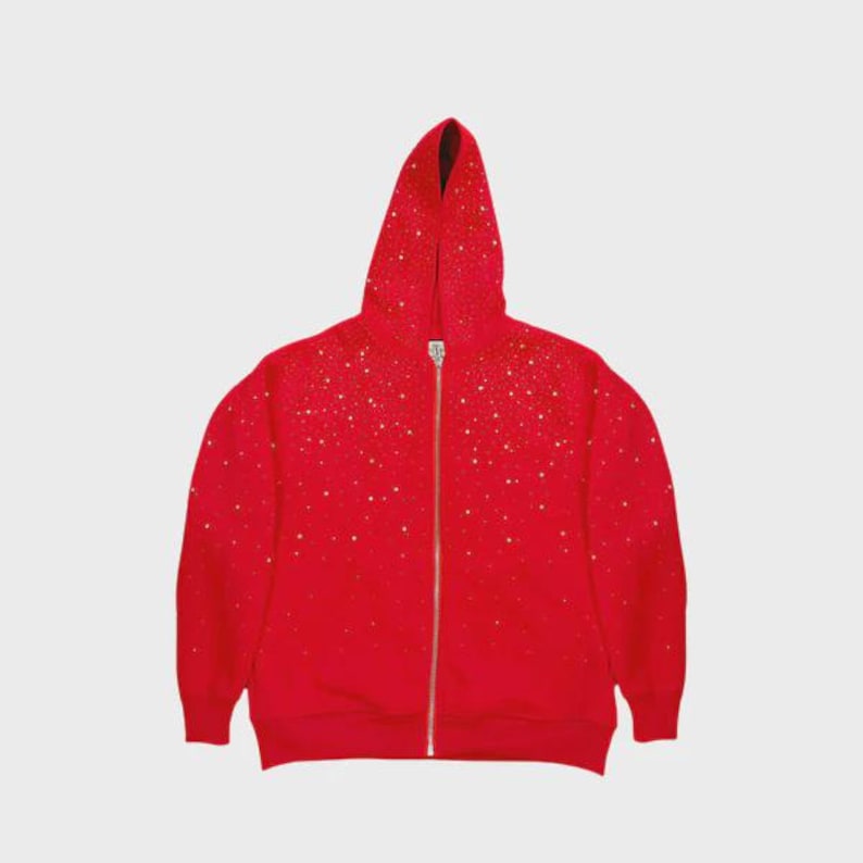 May include: A red zip-up hoodie with a hood, adorned with scattered gold-colored embellishments. The hoodie features a full-length zipper and is made of a soft material. The garment is displayed against a plain white background.